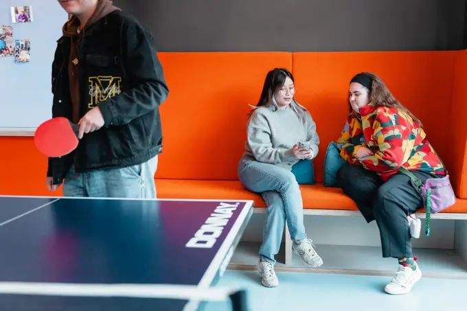 Two students chatting on an orange bench while another student plays table tennis in foreground.